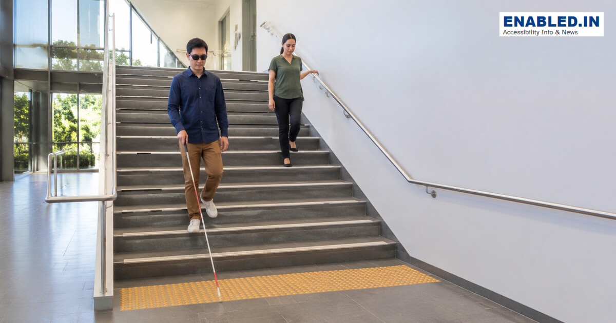 Person using a white cane descending a well-lit stair with continuous handrails, contrasting nosings and a tactile warning surface.